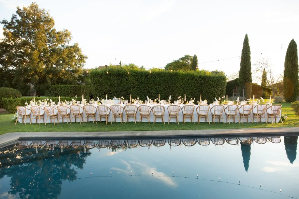 Long outdoor banquet table by a pool, white linens, flowers and cypress trees