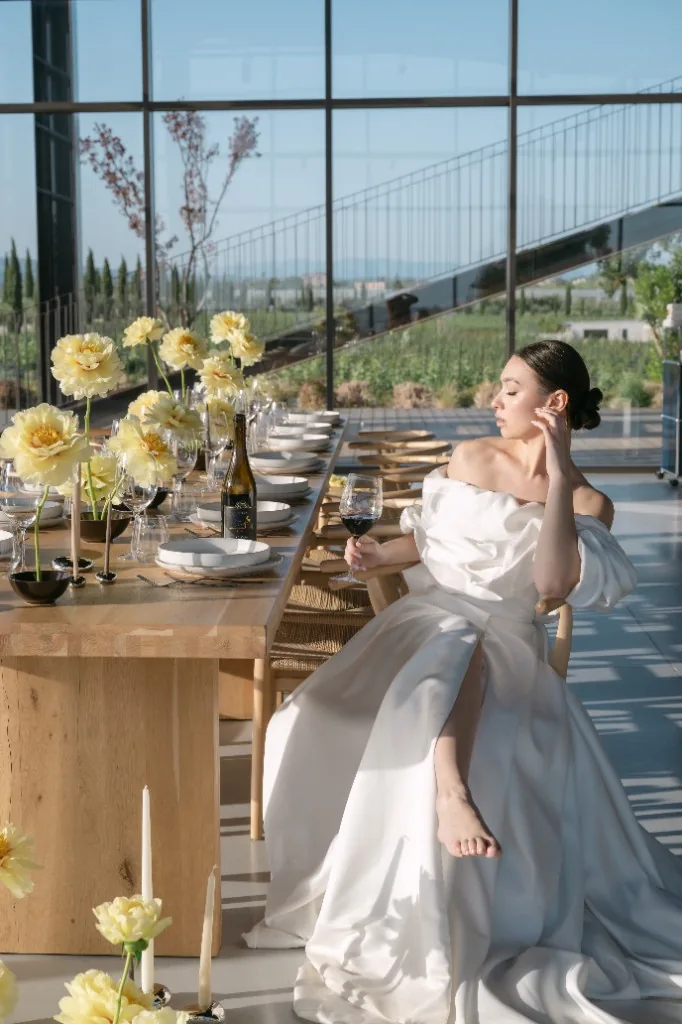 Wedding editorial in Latina: bride in white at a long wooden table with yellow flowers, wine and vineyard views through floor-to-ceiling windows