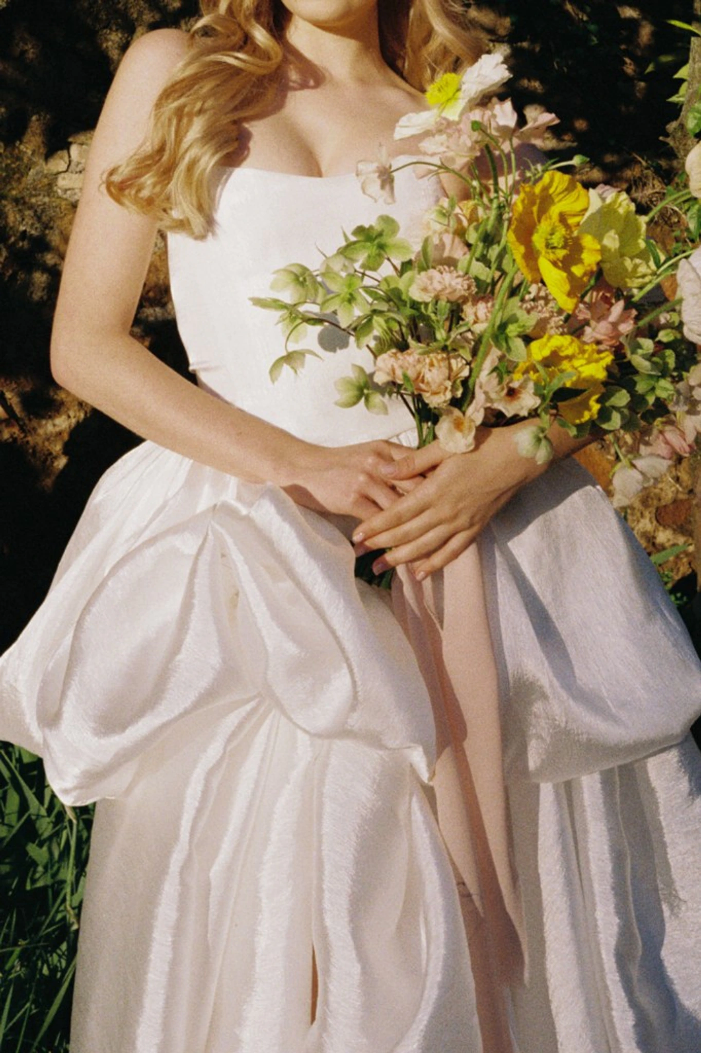 Bride in a textured ivory wedding gown with a lush bouquet of yellow blooms and flowing ribbons, warm sunlight and garden stone wall in the background