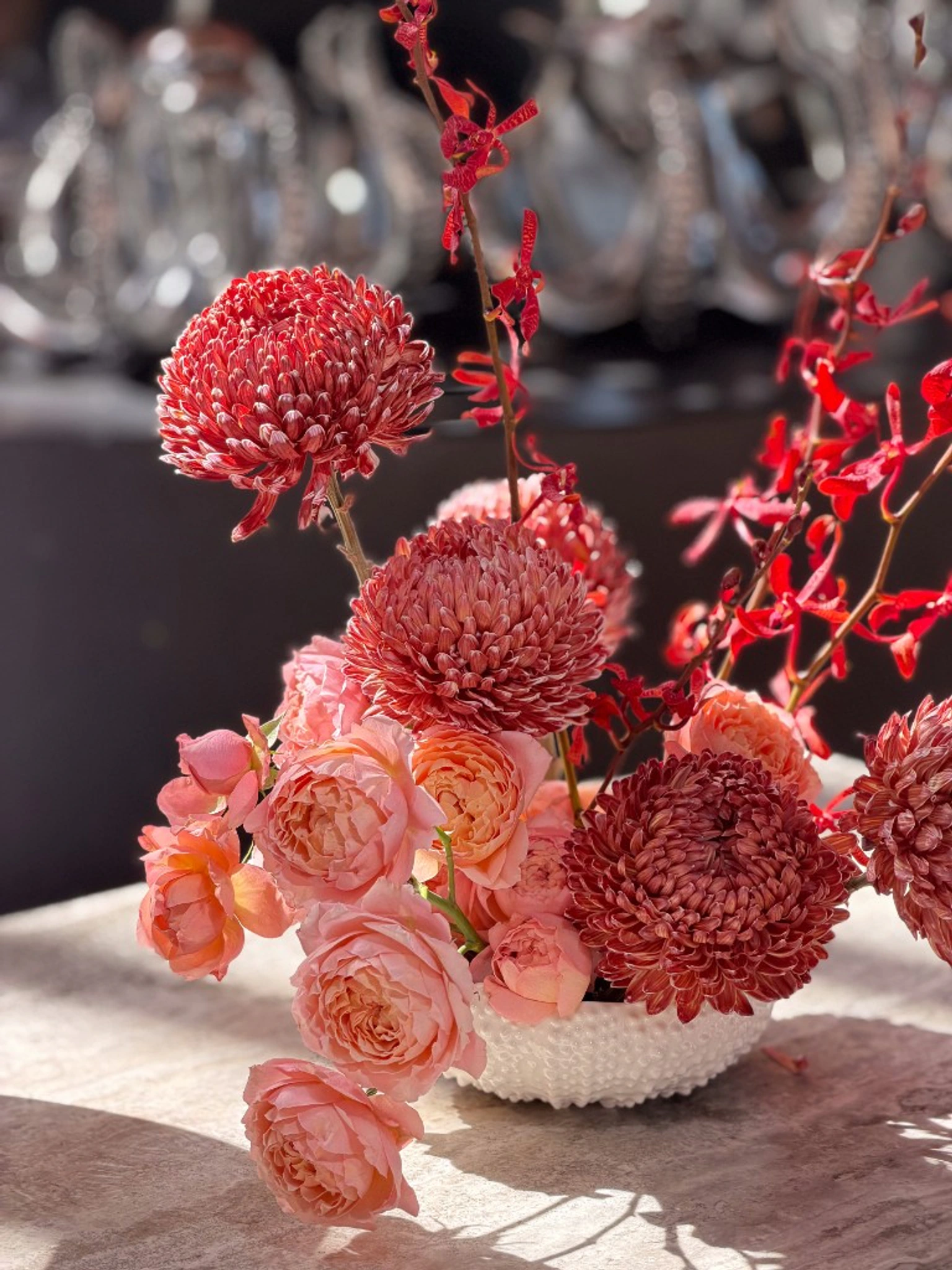 Editorial centerpiece with coral chrysanthemums blush roses red orchids in a sculpted white vase on luminous stone tabletop