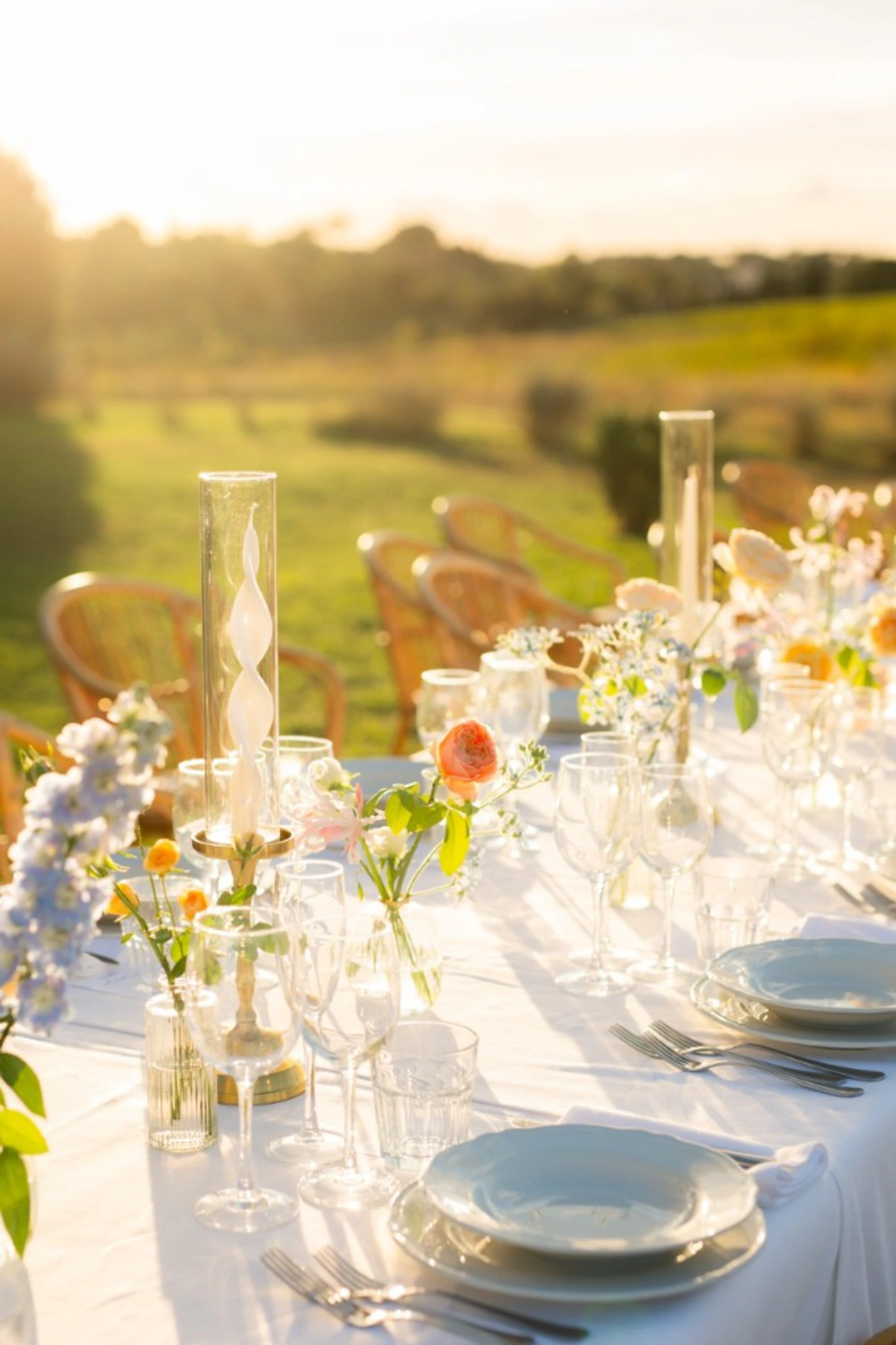Outdoor reception table lined with wicker chairs blush florals and candlelight photographed at golden hour with fields fading into sunset
